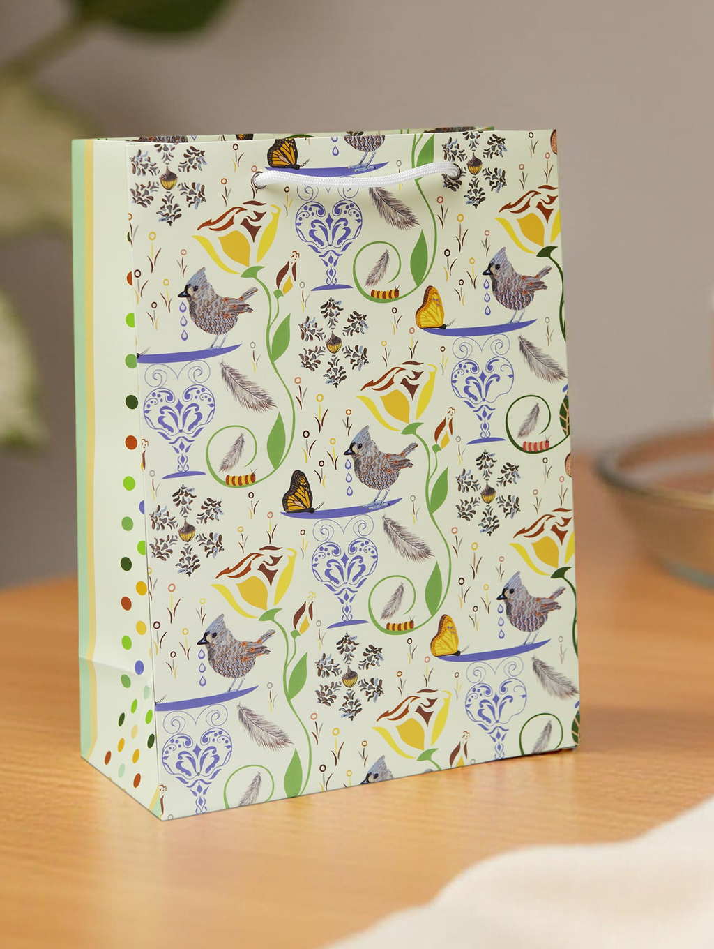 Decorative gift bag with rope handles and sturdy construction. Design has birds, butterflies, flowers, and caterpillars on a pale yellow background. Paper was responsibly sourced.