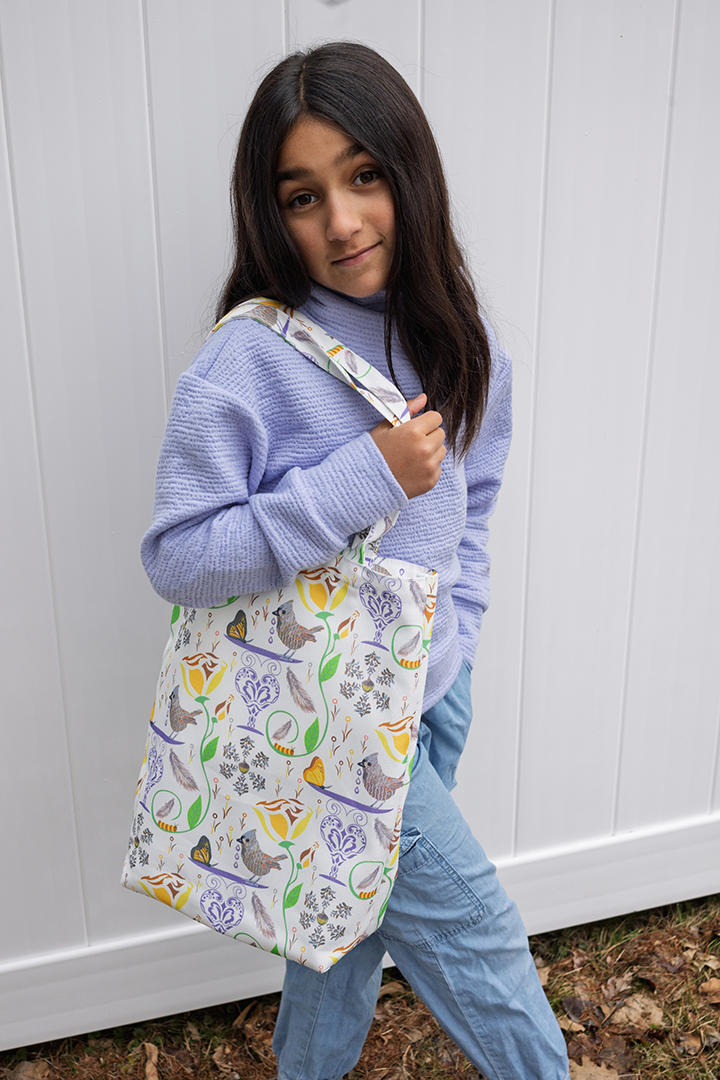 Young lady holding 100% recycled plastic bag with a repeat design of birds, butterfies, flowers, and caterpillars on a white background.