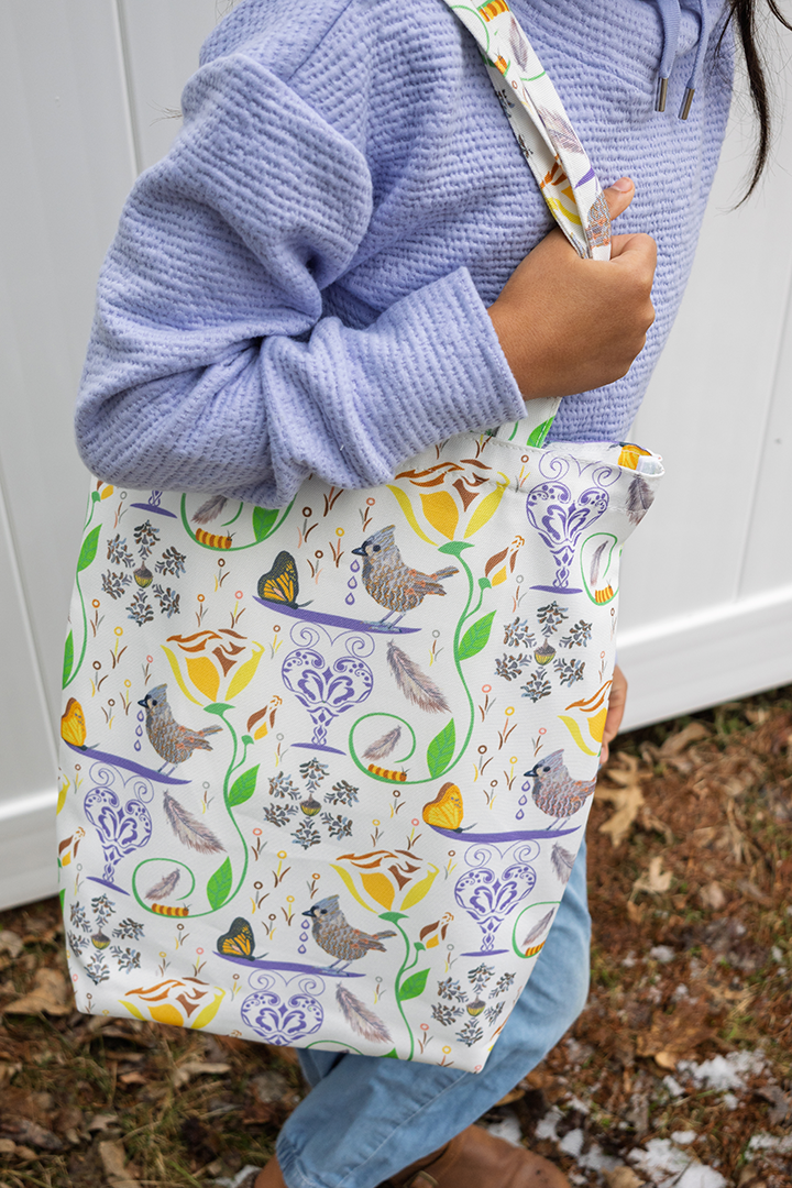Person holding a 100% recycled plastic bag with a repeat design of birds, butterfies, flowers, and caterpillars on a white background.