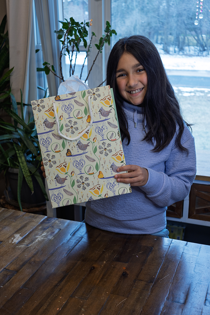 Girl opening gift bag. Decorative gift bag with rope handles and sturdy construction. Design has birds, butterflies, flowers, and caterpillars on a pale yellow background. Tissue is not sold with the bag.