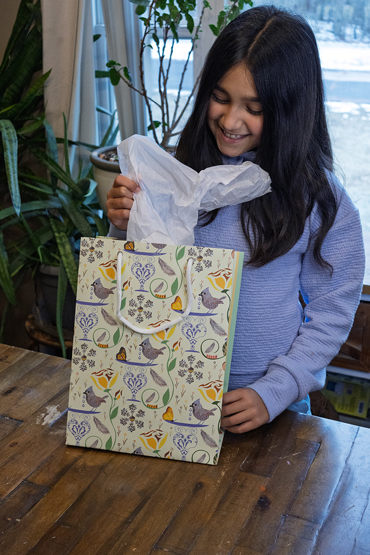 Girl opening gift bag. Decorative gift bag with rope handles and sturdy construction. Design has birds, butterflies, flowers, and caterpillars on a pale yellow background. Tissue is not sold with the bag.