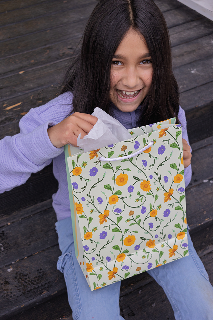 Girl holding a yellow and blue floral gift bag with rope handles. Tissue is not included with the purchase. Bag's paper was responsibly sourced.