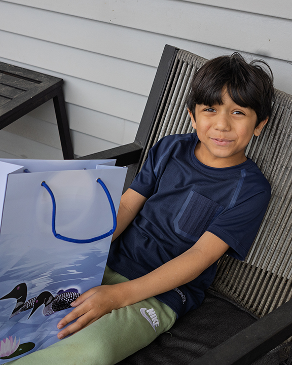 Child sitting on a chair holding a gift bag with a loon design on blue water, outdoors.