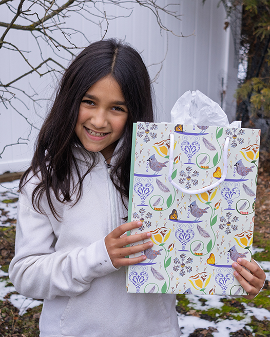 Girl showcasing a gift bag. Girl opening gift bag. Decorative gift bag with rope handles and sturdy construction. Design has birds, butterflies, flowers, and caterpillars on a pale yellow background. Tissue is not sold with the bag.