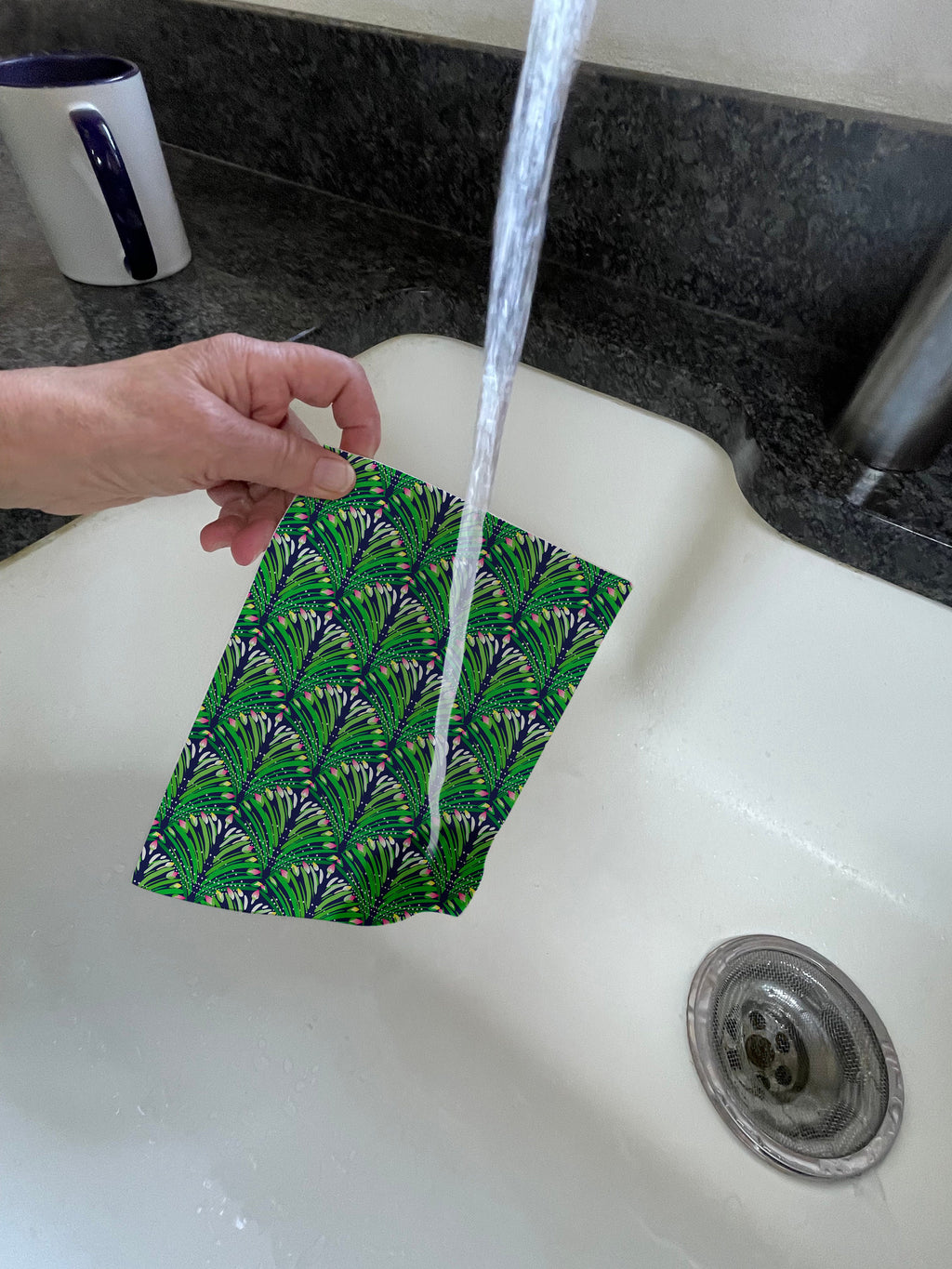 Green, yellow, and pink floral pattern printed on a Swedish dishcloth. Compostable cloth is made from 70% cellulose and 30% repurposed cotton. Dishcloth is being held under running water.