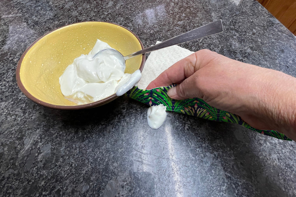 Green, yellow, and pink floral pattern printed on a Swedish dishcloth. Compostable cloth is made from 70% cellulose and 30% repurposed cotton. Person is oping up yogurt with the dishcloth.